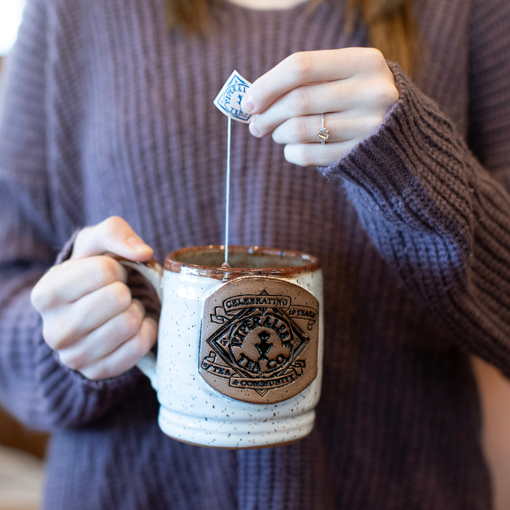 A woman holding a Handmade 10th Anniversary Pottery Mug from Piper & Leaf Tea Co. with a tea bag in it.
