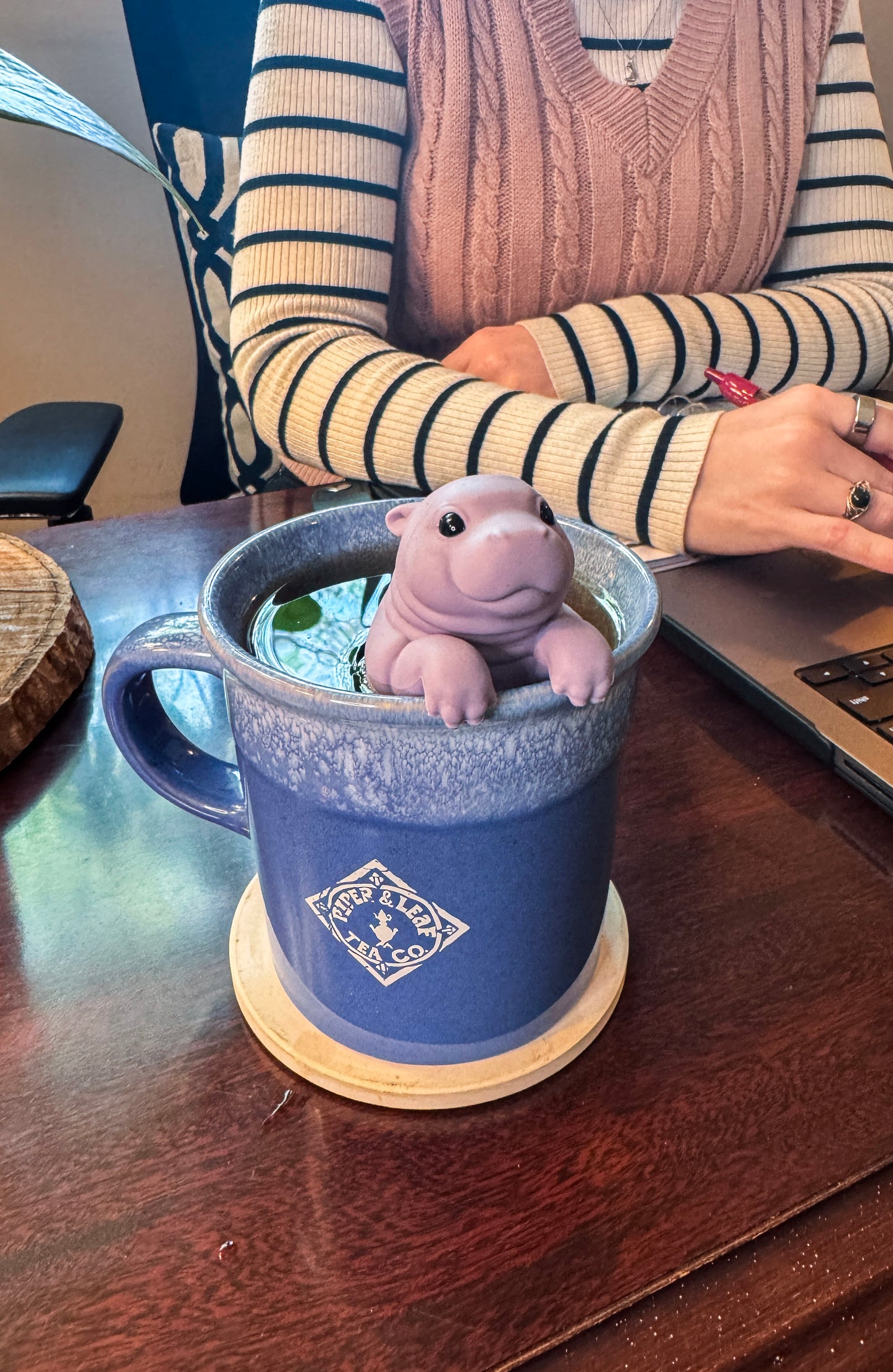 At a desk with a laptop, a Piper & Leaf Tea Co. purple mug holds loose-leaf tea, with the Sip'Opotamus Infuser—a small, realistic hippo—partially submerged in the cup.