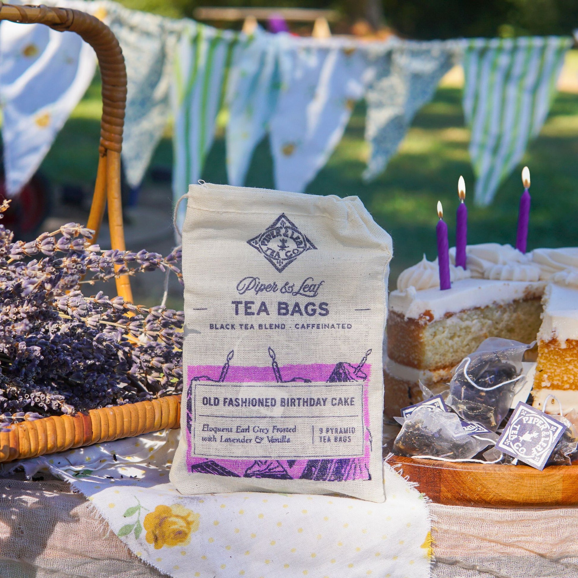 A bag of Piper & Leaf Tea Co.'s Old Fashioned Birthday Cake 9ct Tea Bags in Muslin sits on a table with a slice of cake, tea bags, lavender, and outdoor bunting in the background.