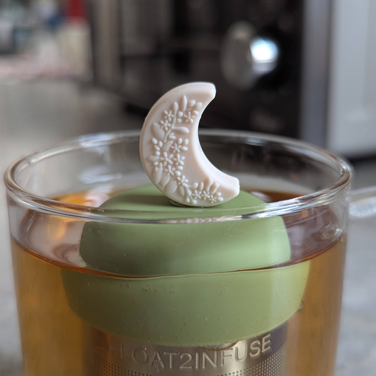 Enjoy your favorite loose-leaf blends with the Ivory Floral Crescent Moon – Floating Tea Infuser by Float2infuse. This elegant glass of tea features a green lid topped with a white, crescent moon-shaped decoration adorned with a floral pattern, complemented by a floating tea infuser for effortless brewing.