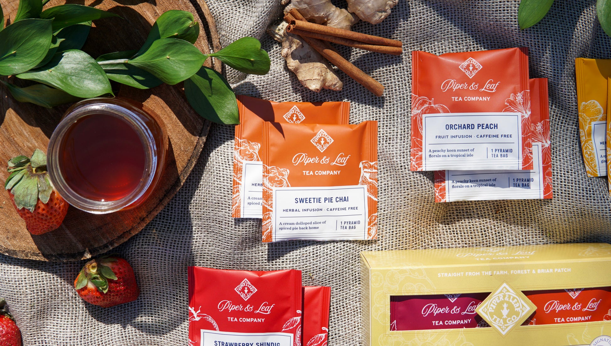 Flat lay of Piper & Leaf Tea Co.’s Caffeine Free Sampler—Box of 16 Tea Bags—with a glass of tea, fresh strawberries, leafy greens, cinnamon sticks, and a wooden tray arranged on a fabric surface.