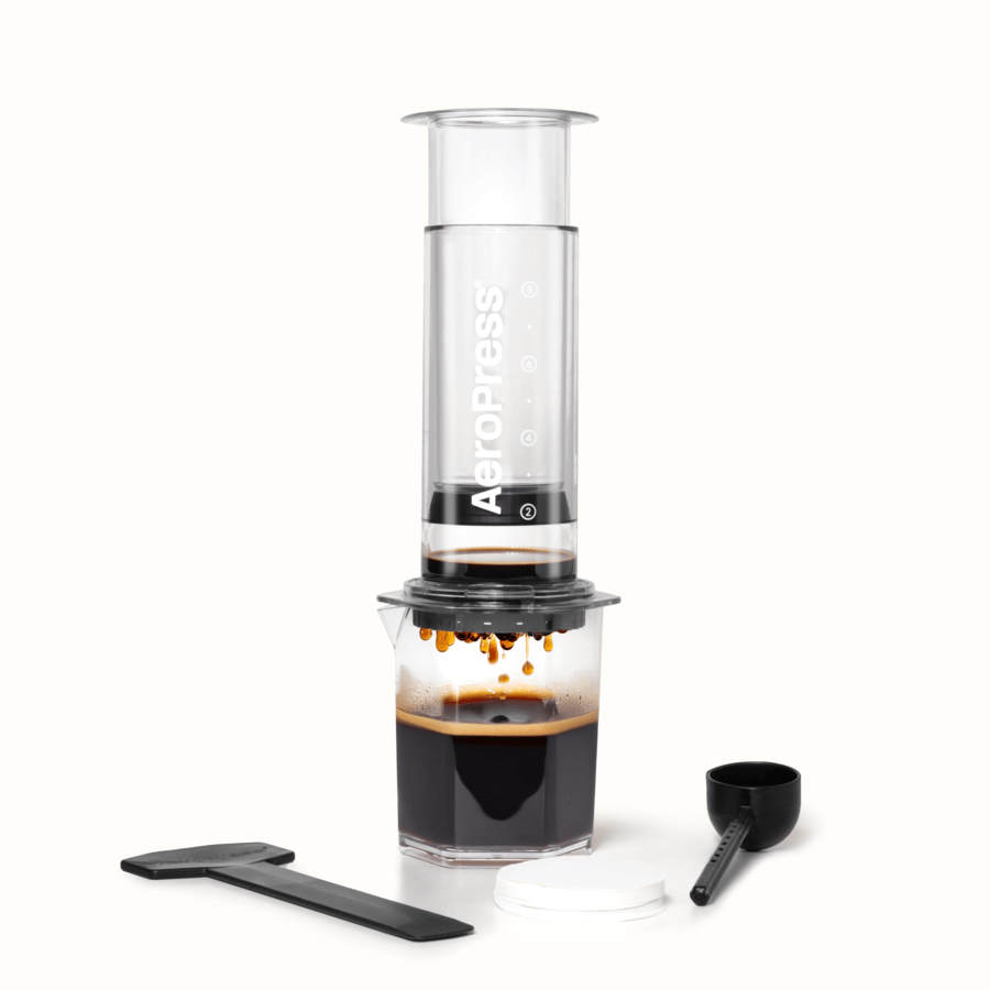 The AeroPress Clear XL Portable Coffee Press by AeroPress brews coffee into a glass, with a stirrer, scoop, and filter papers displayed in front—demonstrating the versatility of this extra large portable coffee maker.