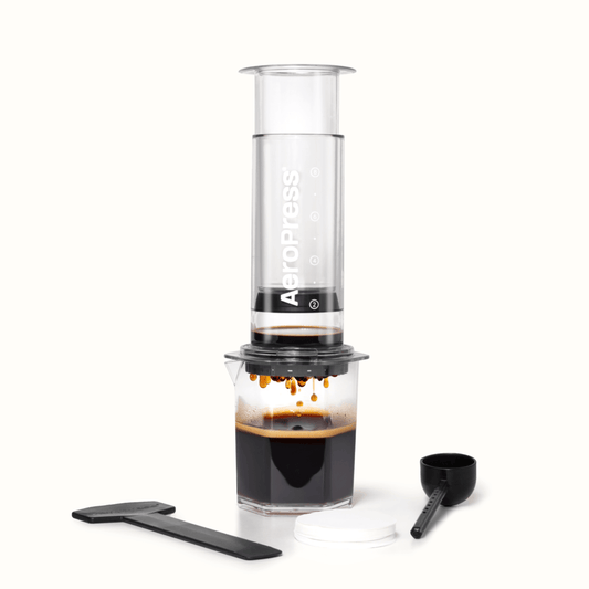 The AeroPress Clear XL Portable Coffee Press by AeroPress brews coffee into a glass, with a stirrer, scoop, and filter papers displayed in front—demonstrating the versatility of this extra large portable coffee maker.