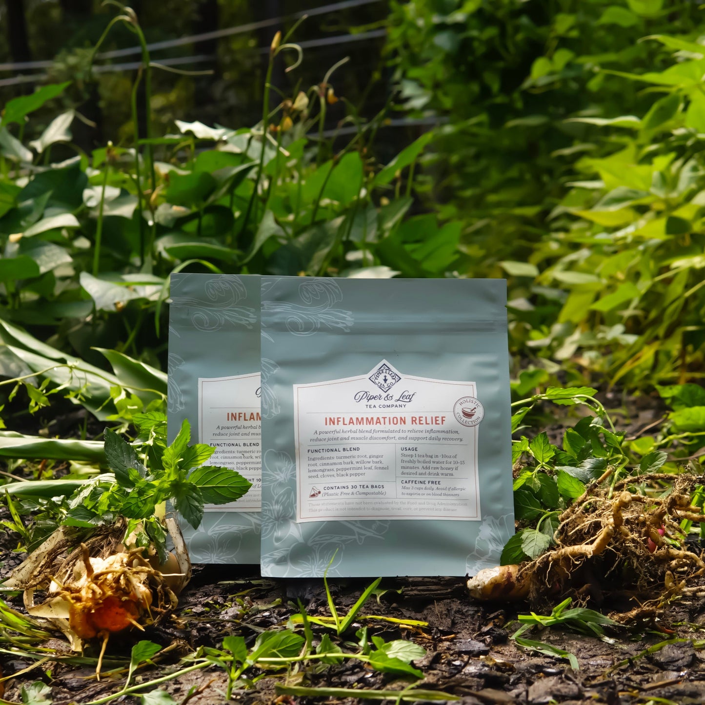 Two upright packages of Piper and Leaf Tea Co.’s Inflammation Relief rest among green plants and wilted roots, featuring a natural herbal blend formulated to help soothe inflammation and provide relief for joint and muscle discomfort.