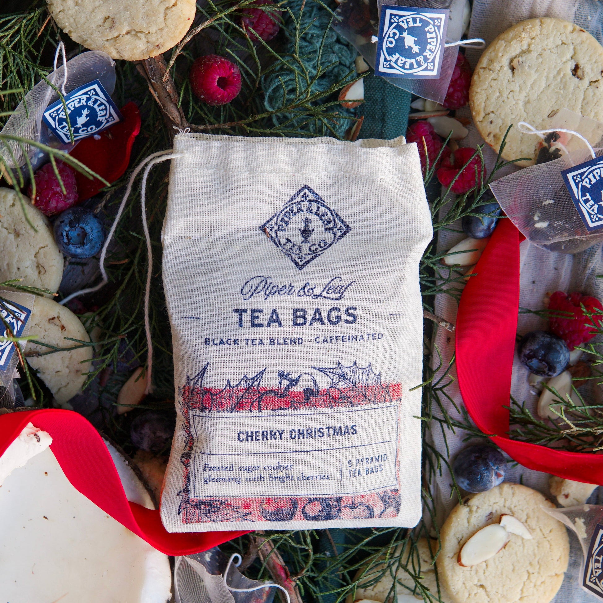 A bag of Piper & Leaf Tea Co. Cherry Christmas 9ct Tea Bags in Muslin sits among cookies, loose berries, pine needles, and red ribbon.