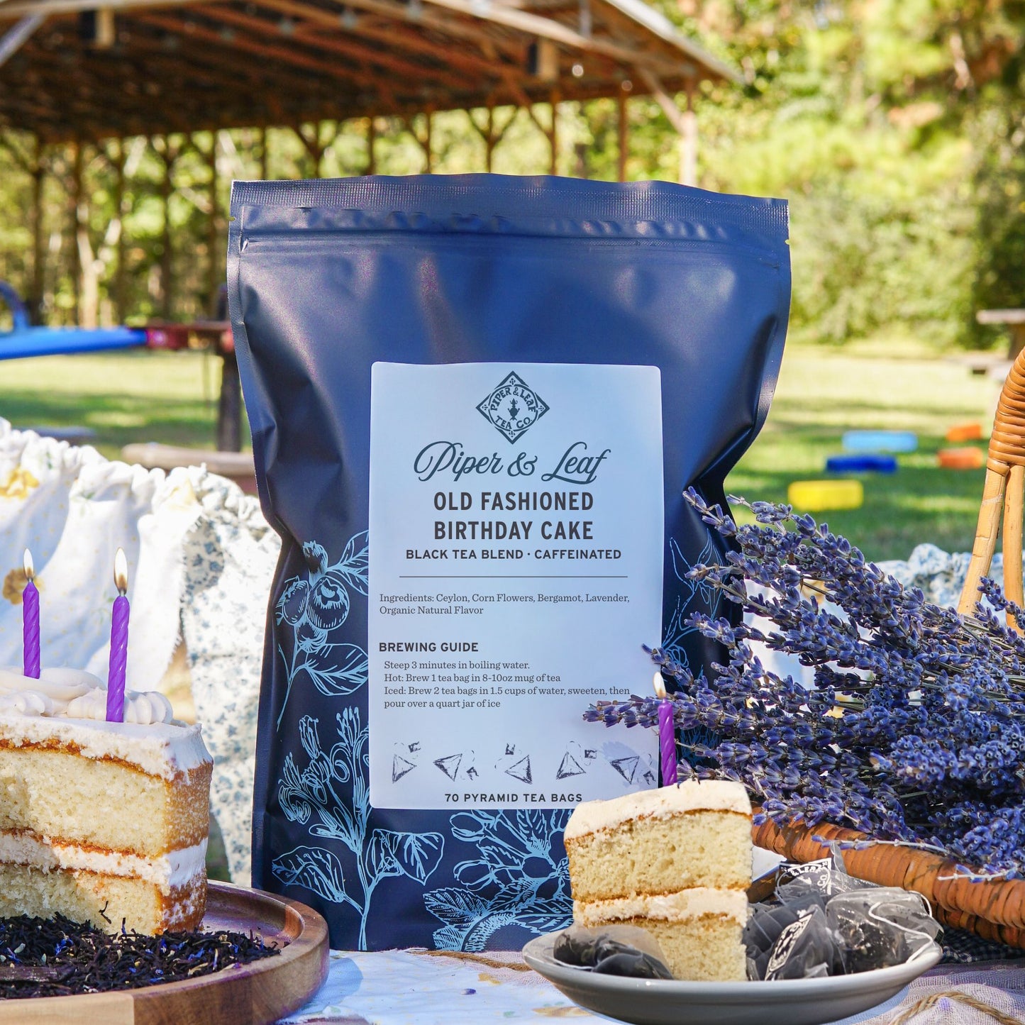 A bag of Piper & Leaf Tea Co. Old Fashioned Birthday Cake Bulk Sachets - 70ct Tea Bags rests on a picnic table with birthday cake slices, a lavender basket, and festive outdoor decorations in the background.