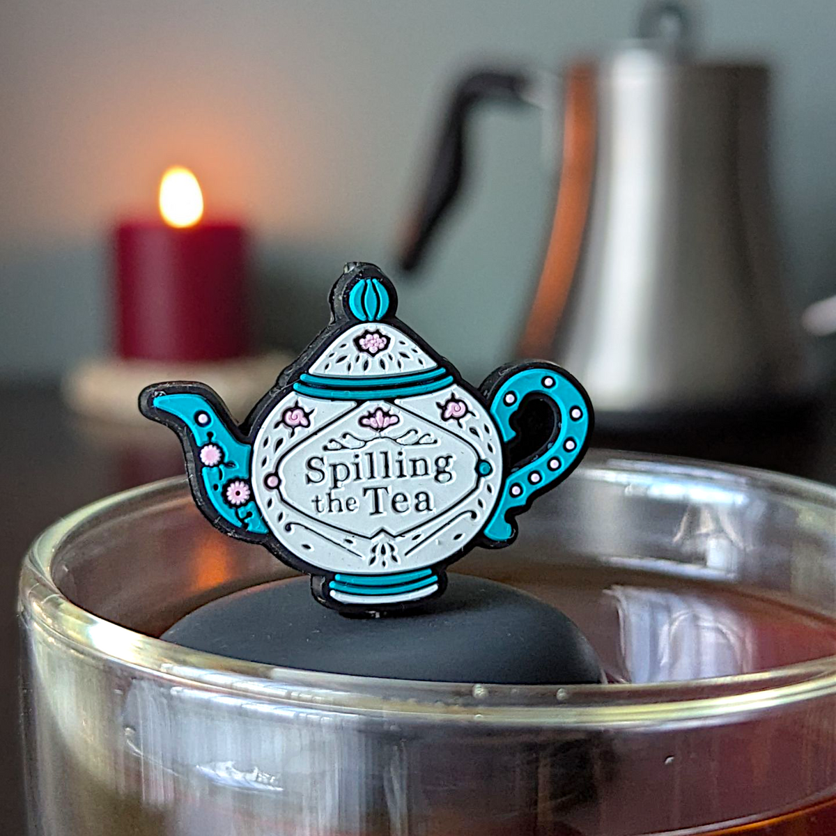 The Spill the Tea - Floating Tea Infuser by Float2infuse Tea Infuser, designed in the shape of a teapot emblazoned with “Spilling the Tea,” floats atop a glass of tea as a whimsical and charming accessory, with a glowing candle and sleek metal kettle softly blurred in the background.