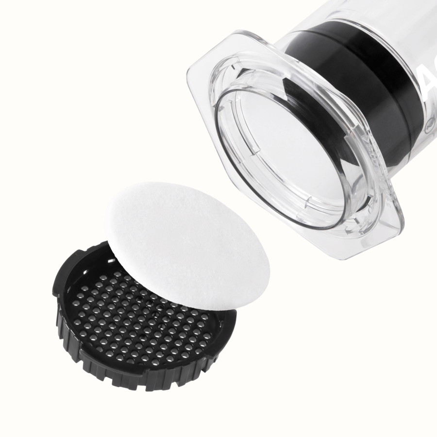 A close-up of the AeroPress Clear XL Portable Coffee Press by AeroPress highlights its transparent cylindrical design, black filter cap, and a circular white pad beside the cap—ideal for anyone wanting an extra-large portable coffee brewer.