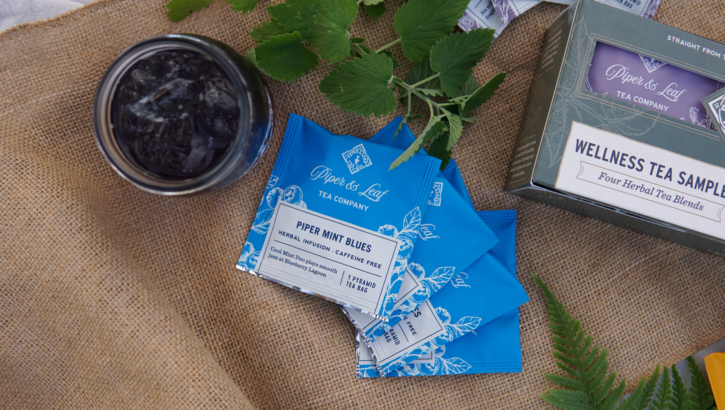 Packets of Piper & Leaf Tea Co. "Piper Mint Blues" caffeine-free tea, a glass of iced tea with mint leaves, and a Wellness Tea Sampler - Box of 16 Tea Bags are artfully arranged on burlap.