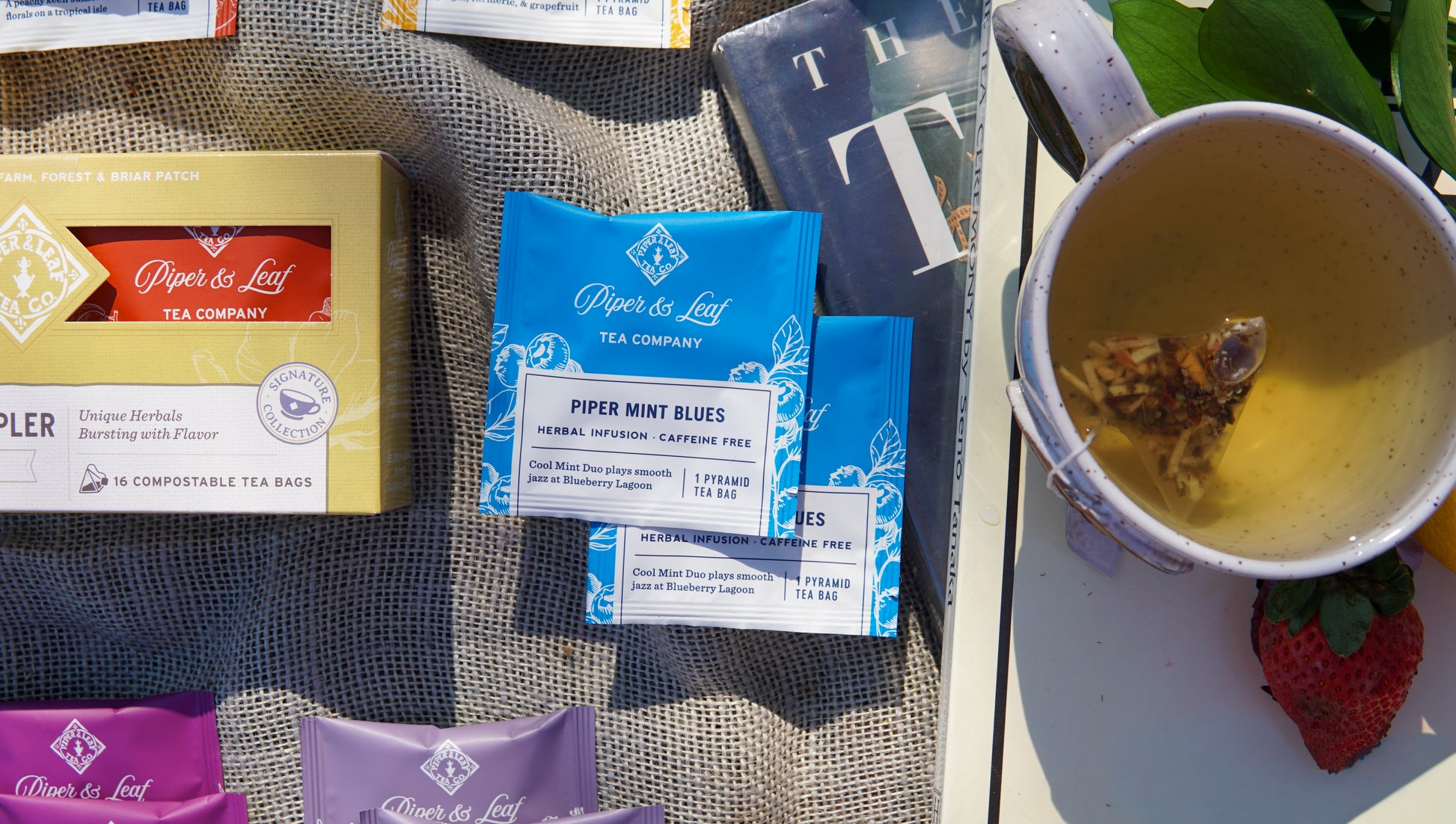 A Piper & Leaf Tea Co. Caffeine Free Sampler (Box of 16 Tea Bags) is displayed on a cloth beside a cup of brewed tea, a strawberry, and an open book.