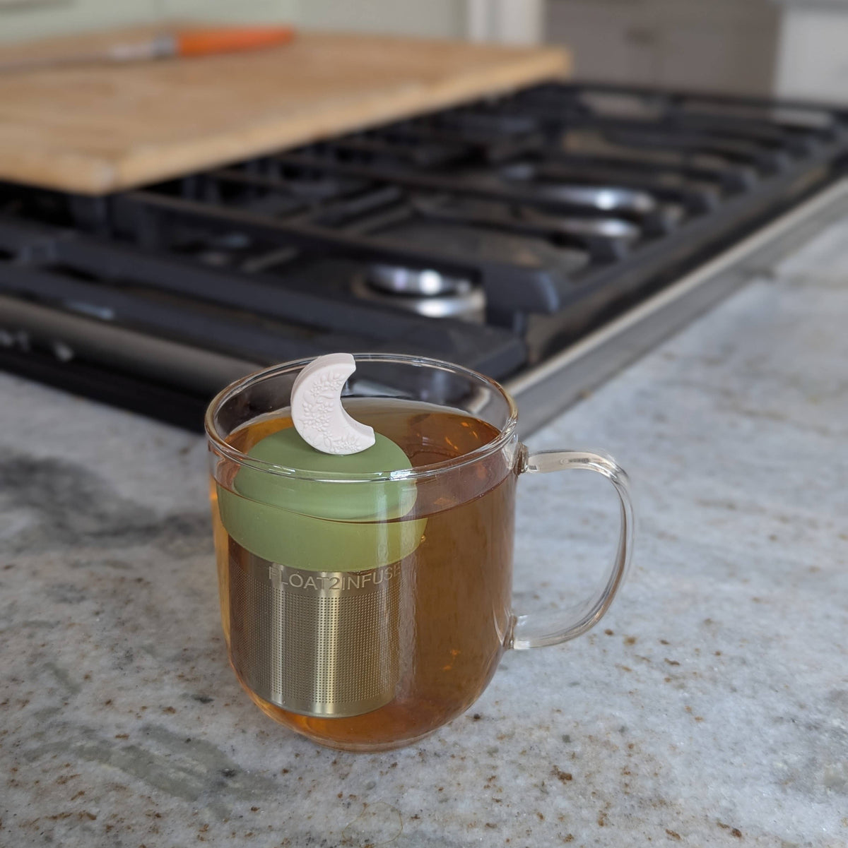A glass mug of tea rests on a kitchen counter, featuring the Float2infuse Tea Infuser’s Ivory Floral Crescent Moon – a floating loose-leaf tea infuser with a green silicone lid and a distinctive white crescent-shaped handle. This chic and functional accessory adds style to your tea ritual, while a stovetop and wooden cutting board enhance the cozy atmosphere of the kitchen in the background.