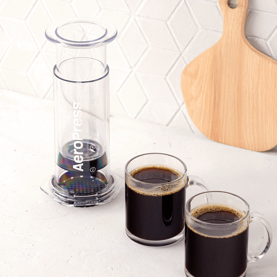 An AeroPress Clear XL Portable Coffee Press by AeroPress and two glass mugs of black coffee sit on a white countertop, with a wooden cutting board in the background.