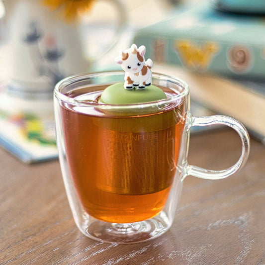 A glass cup of tea sits on the table with the Float2infuse Tea Infuser—Baby Goat floating inside. The infuser’s lid is topped with an adorable baby goat figurine, adding a whimsical touch to this charming tea accessory. Books can be seen softly blurred in the background.