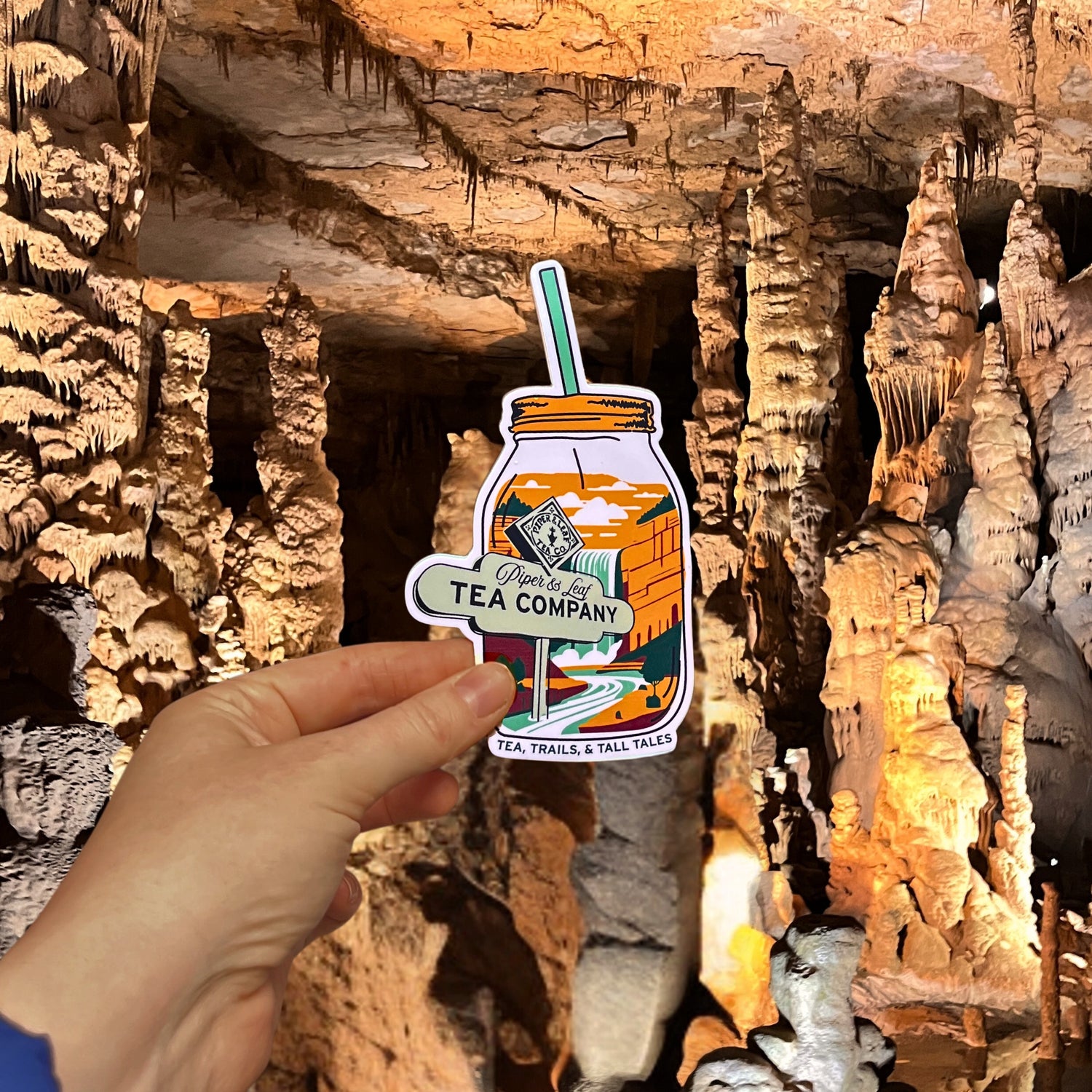 A hand holds a "Tea, Trails & Tall Tales Adventurer Sticker" by Piper & Leaf Tea Co., shaped like a tea jar with cave stalactites and stalagmites in the background, ideal for adventure lovers and perfect for decorating travel mugs.