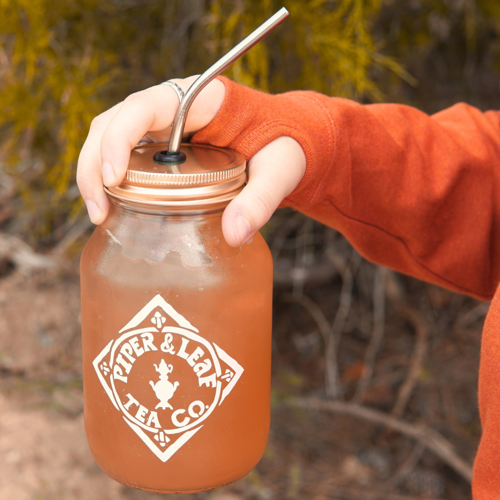 A person in an Autumn Leaf Hoodie holds a large glass jar with a metal lid and straw, containing an iced beverage from Piper & Leaf Tea Co.