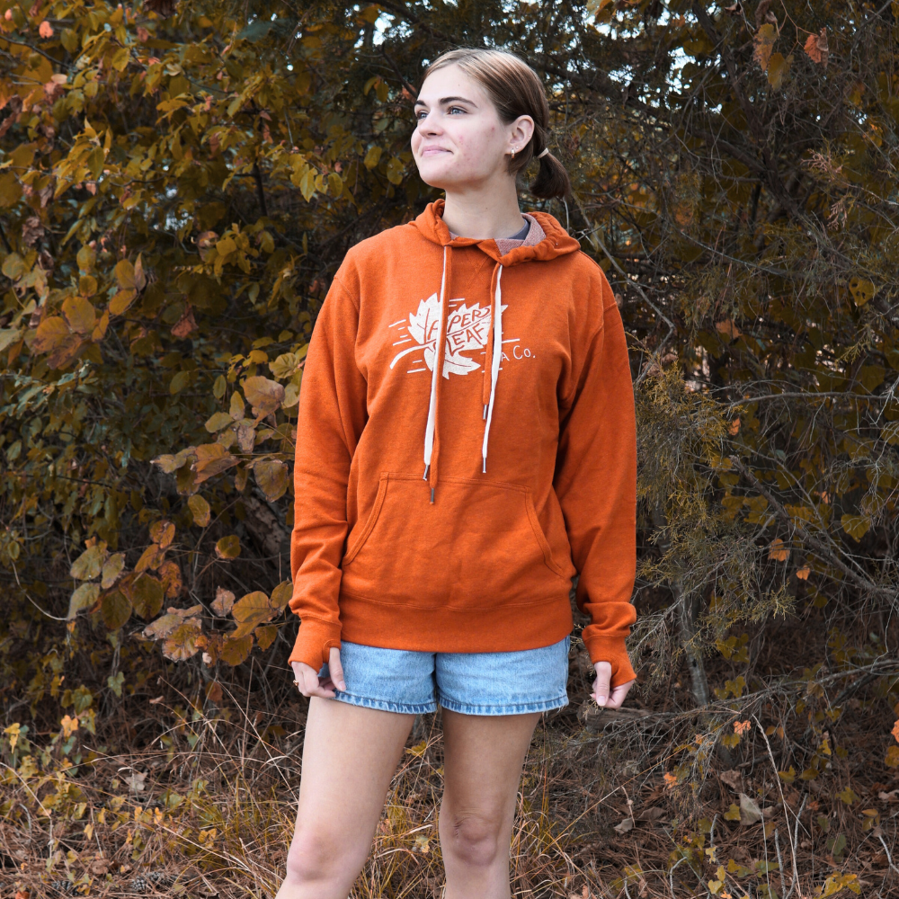 A person wearing a Piper & Leaf Tea Co. Autumn Leaf Hoodie and denim shorts stands outdoors against a background of trees and foliage.