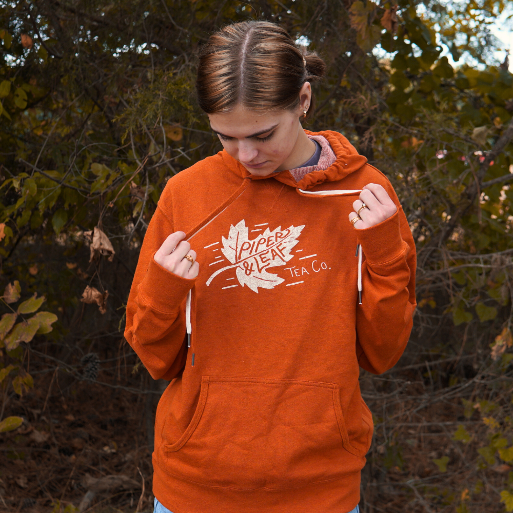 Person wearing an Autumn Leaf Hoodie from Piper & Leaf Tea Co., looking down and adjusting the hood strings, standing outdoors with foliage in the background.