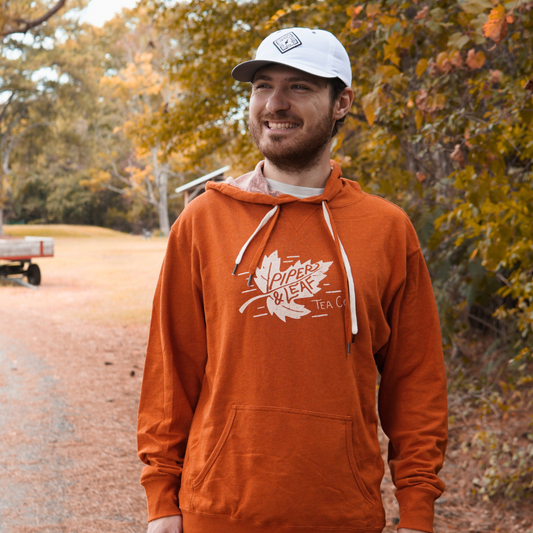 A man in a Piper & Leaf Tea Co. Autumn Leaf Hoodie and white cap stands outdoors on a leaf-covered path, surrounded by autumn foliage.