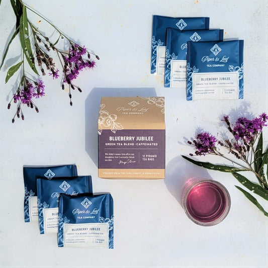 A box of Blueberry Jubilee- Box of 12 Individually Wrapped Tea Bag Sachets from Piper & Leaf Tea Co. and a glass of tea next to purple flowers.