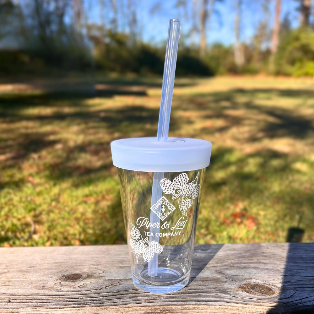 A Limited Edition: Into the Briar Patch P&L X Silipint 16oz Cup, made of unbreakable clear plastic and complete with a straw, rests on a wooden surface. Adorned with the Piper & Leaf Tea Co. logo, this BPA-free cup is perfect for outdoor settings against a backdrop of lush green grass and trees under a clear sky.