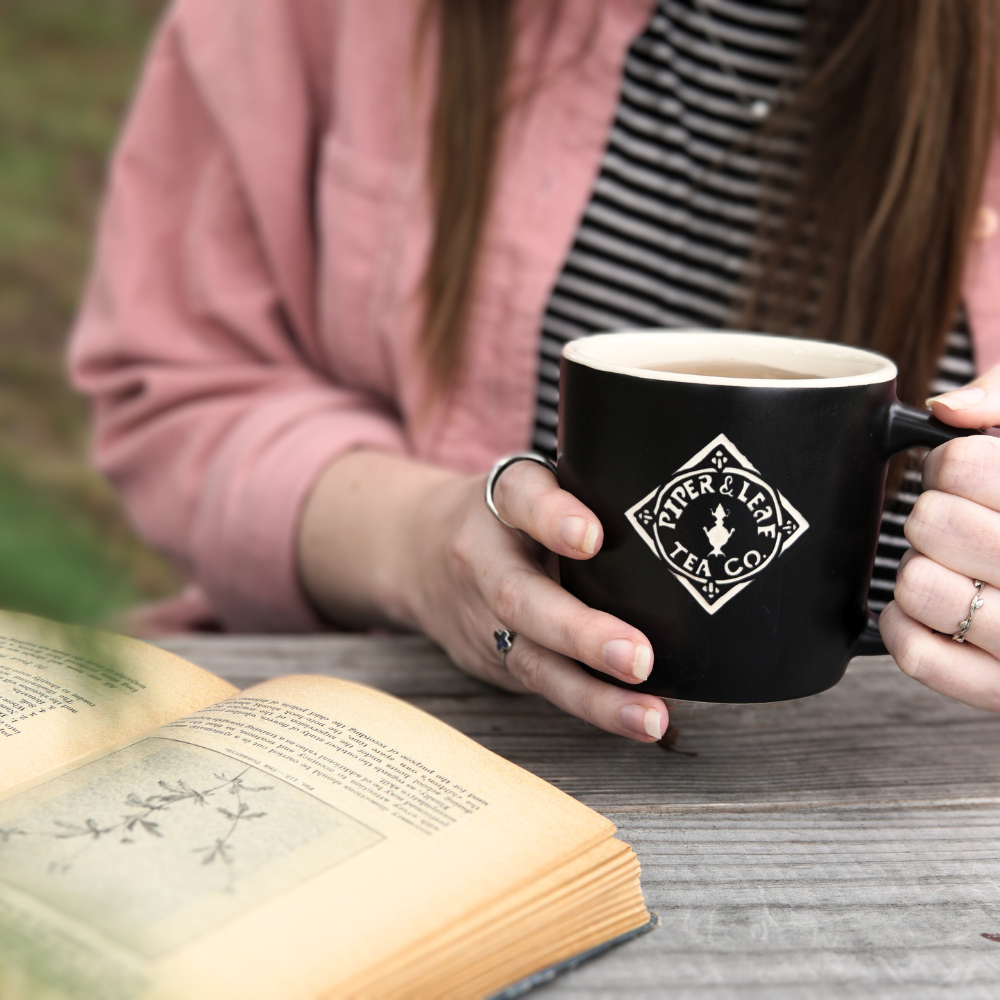 A person holding the 12oz Sand Carved Logo Mug in Matte Black from Piper & Leaf Tea Co. sits at a table with an open book.