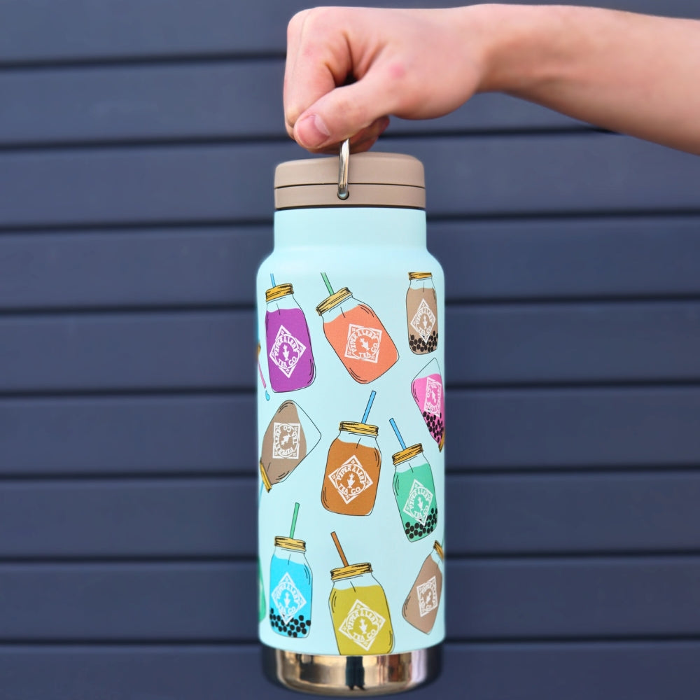 A person holds a 32oz Klean Kanteen by Piper & Leaf Tea Co., featuring colorful boba tea illustrations on light blue. This BPA-free stainless steel tumbler, set against a dark striped background, boasts double-walled vacuum insulation for perfect drink temperature control.