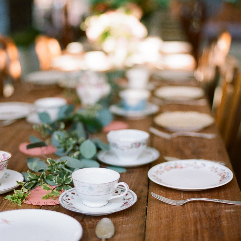 Elegant outdoor dining setting featuring Springtime Afternoon Tea Party, vintage china, silverware, and leafy green decorations on a long wooden table by Piper and Leaf Tea Co.