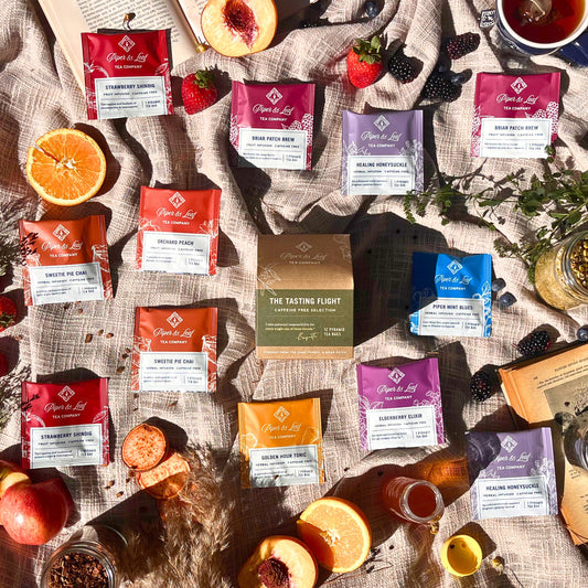 A flat lay featuring Piper & Leaf Tea Co.'s "Caffeine Free Tasting Flight" box of 12 tea bags, artfully arranged on a cloth backdrop with an array of fruits, a candle, books, and scattered loose tea leaves, offering a delightful sensory experience.