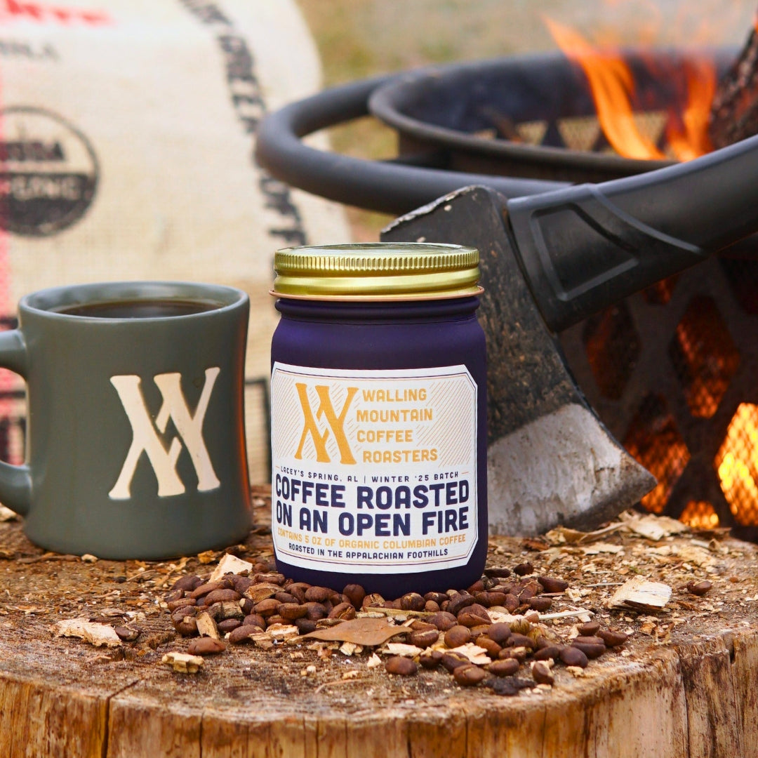 A 5oz jar of Walling Mtn.'s Campfire Roast Coffee sits alongside a mug on a tree stump near a fire pit, with roasted beans scattered around. An axe is lodged in the stump, echoing traditional roasting methods.