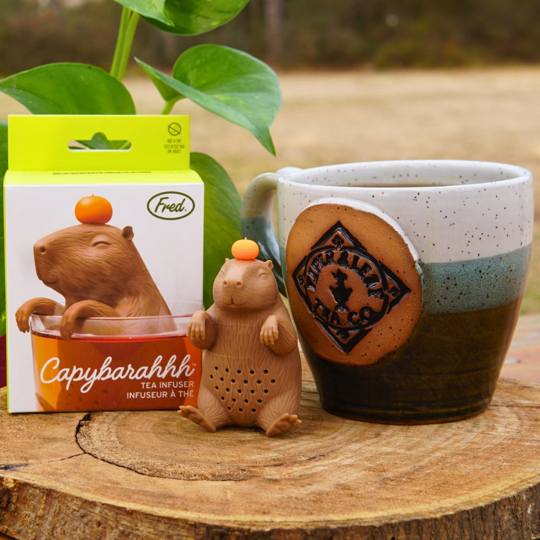 The Capybarahhh Tea Infuser by Piper & Leaf Tea Co. sits beside its packaging and a ceramic mug, with plant leaves in the backdrop.