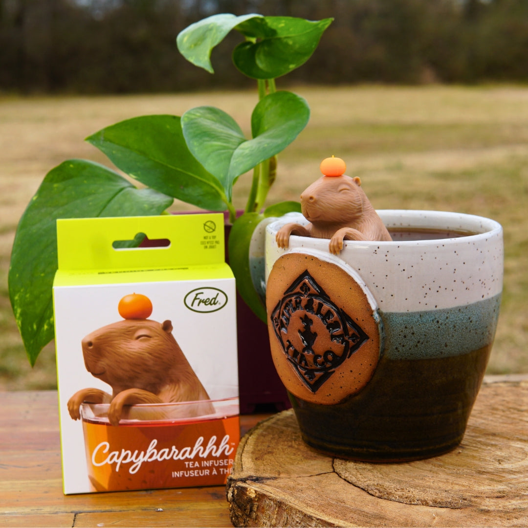 A Capybarahhh Tea Infuser by Piper & Leaf Tea Co. sits in a mug, next to its packaging.