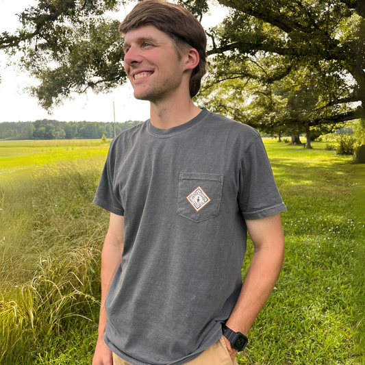 A man stands on grass near trees, smiling in a relaxed-fit Piper & Leaf Tea Co. Crafted in Alabama Tee (100% cotton, gray, pocket) and tan pants.