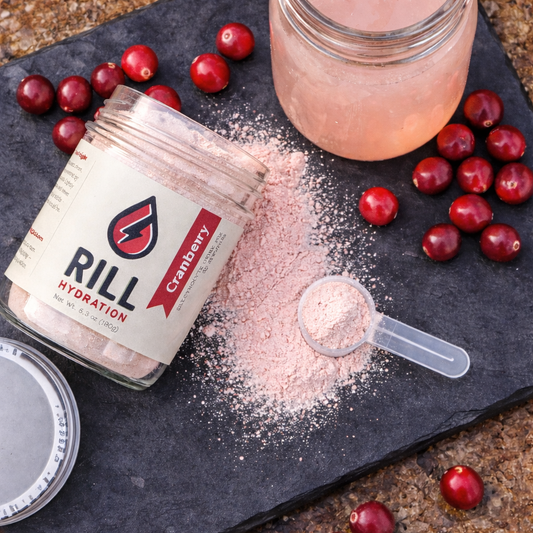 A jar of Rill Hydration's Cranberry Rill Clean Hydration Drink Mix is spilled on slate, with a scoop of powder, fresh cranberries, and a glass of pink drink nearby. Bulk jar contains 30 servings.