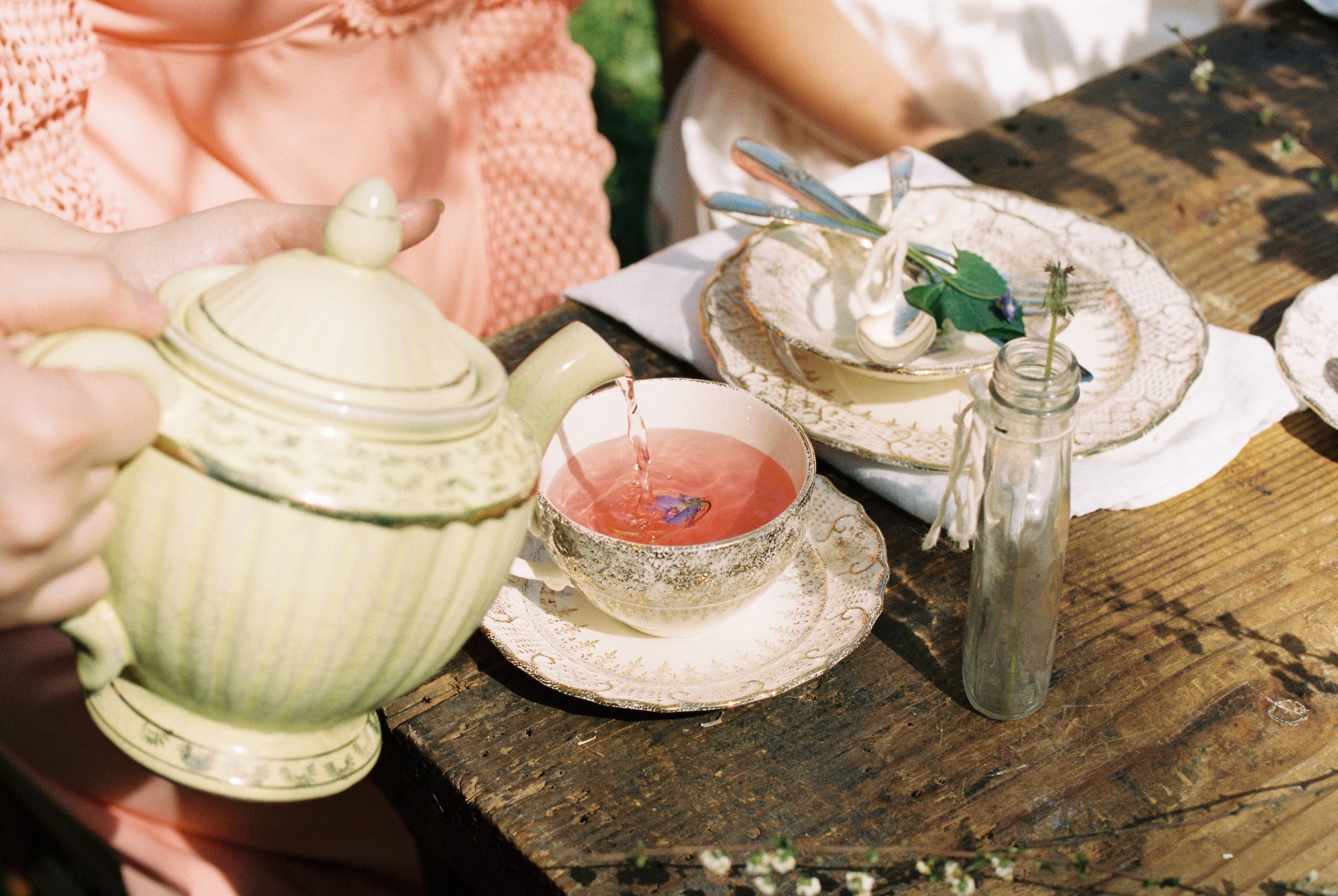 A person pours Afternoon Tea At Piper & Leaf from a green teapot into a floral teacup on a rustic wooden table set with plates, cutlery, and a small flower vase during an inviting afternoon tea.