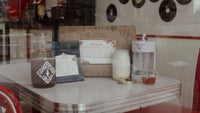 The Piper and Leaf Tea Co. Root Brew Kit includes instructions, tea bags, glass jars with concentrated tea drinks, and a measuring cup, all displayed on a white retro diner table next to a red booth.