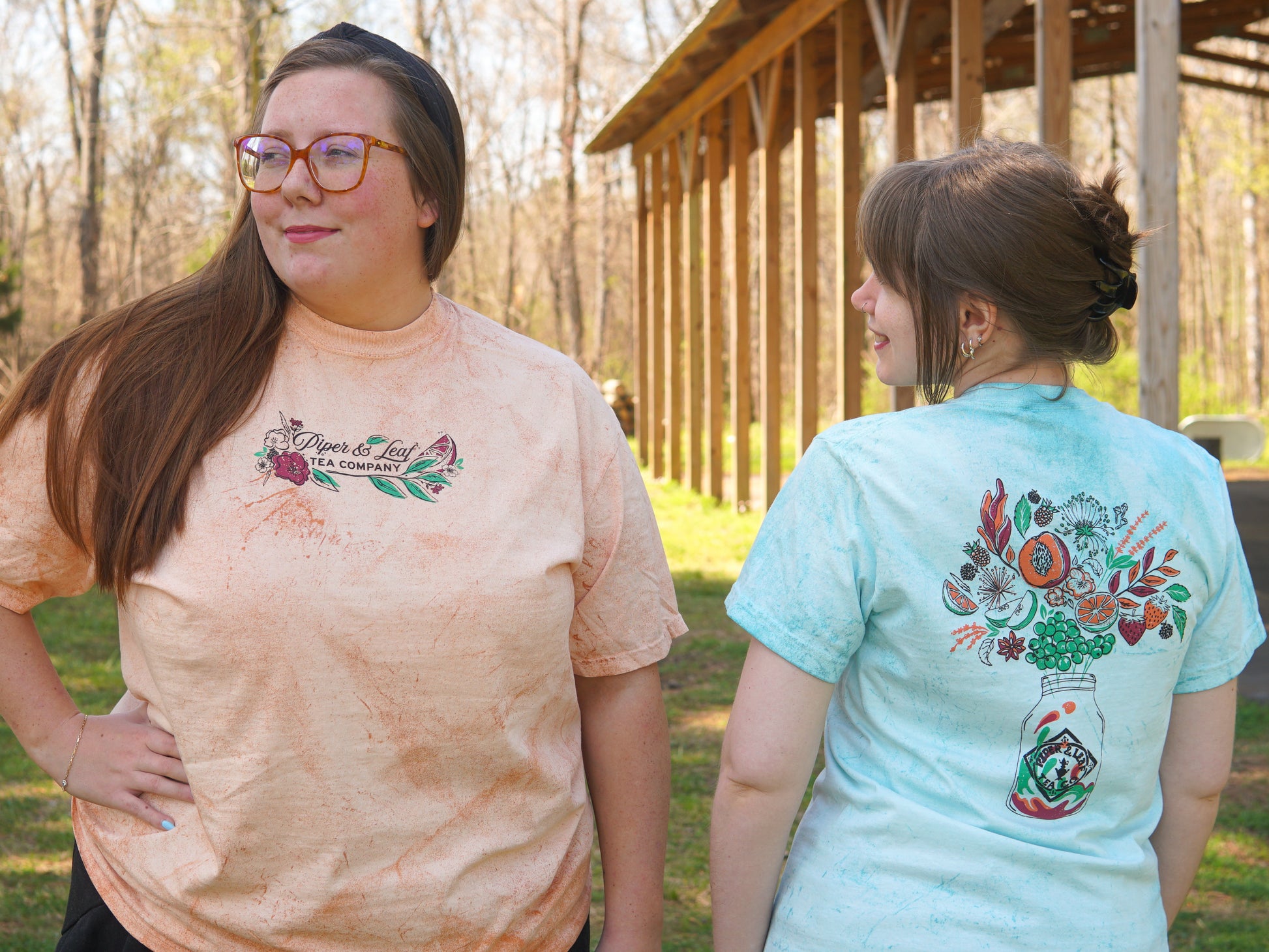 Two women stand outside by a wooden pavilion, each wearing a Piper and Leaf Tea Co. Berries in Bloom Short Sleeve T-shirt featuring floral and tea-inspired designs printed on both the front and back.