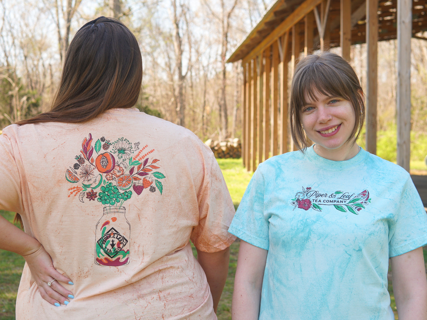 Two women stand outdoors; one faces forward showing a "Piper and Leaf Tea Co." logo on her shirt, while the other reveals a colorful "Berries in Bloom Short Sleeve T-shirt" floral graphic on the back.