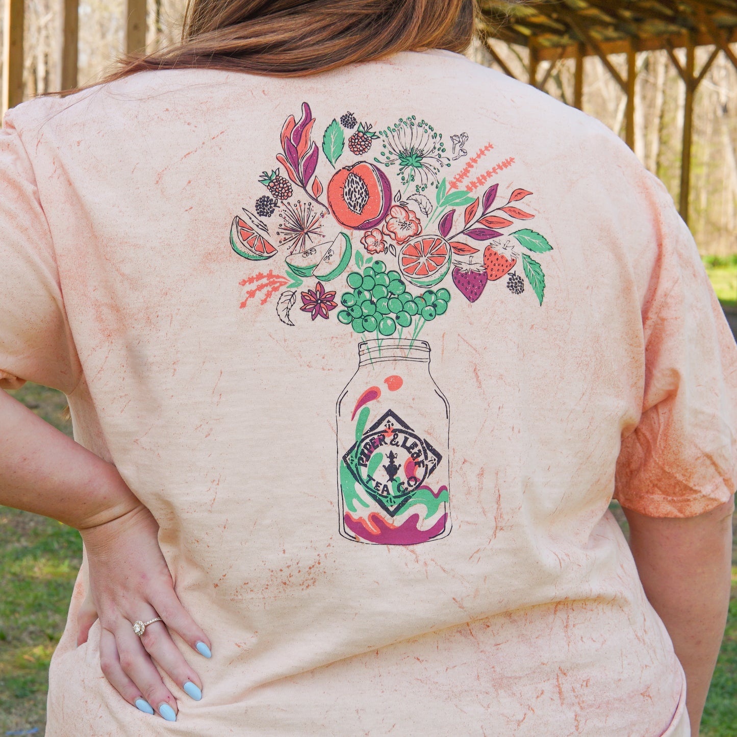 A person stands outdoors wearing the Piper and Leaf Tea Co. "Berries in Bloom" Short Sleeve T-shirt, featuring a colorful fruit and flower mason jar illustration on the back.