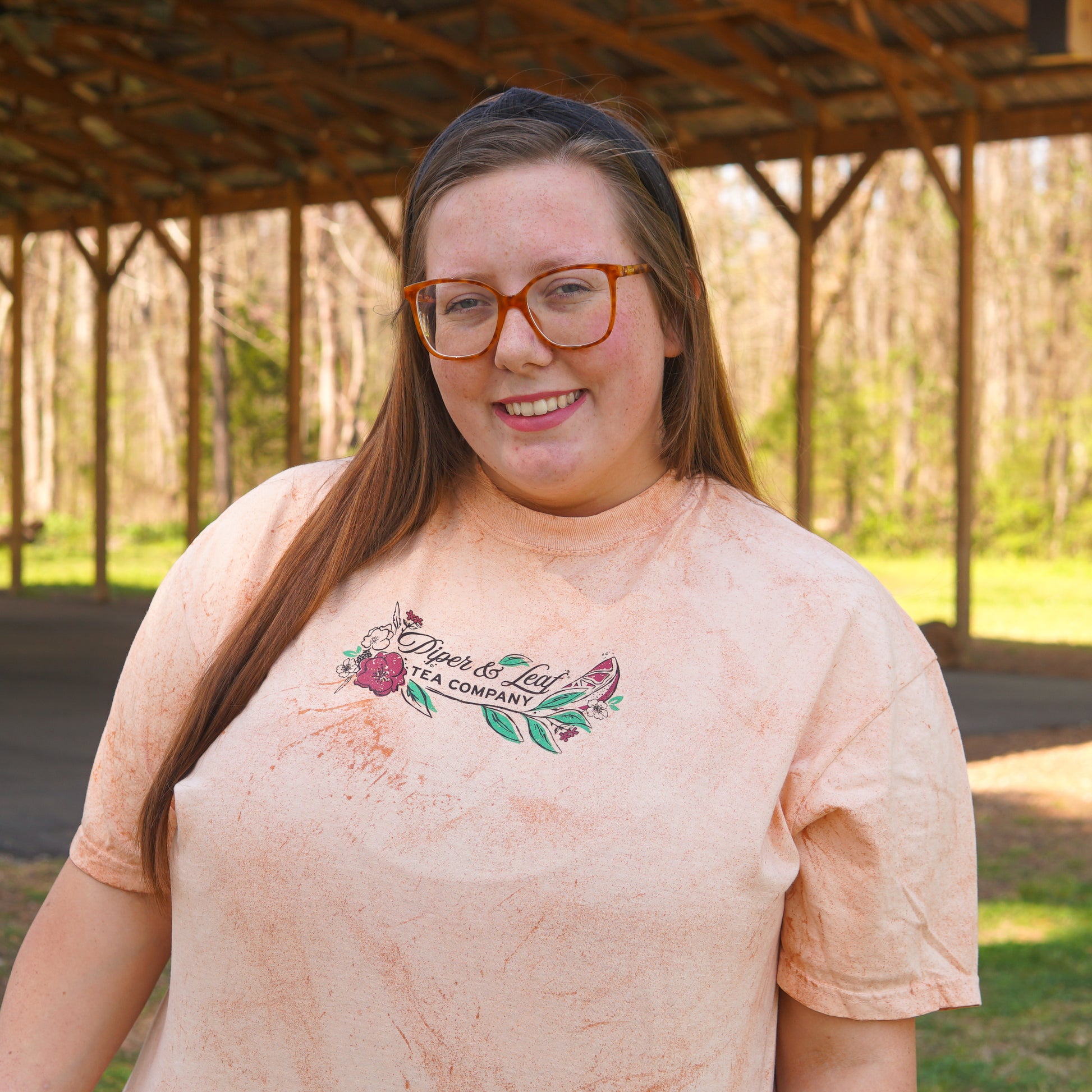 A woman with long brown hair and glasses, wearing a light-colored Berries in Bloom Short Sleeve T-shirt by Piper and Leaf Tea Co., stands outdoors under a wooden shelter.