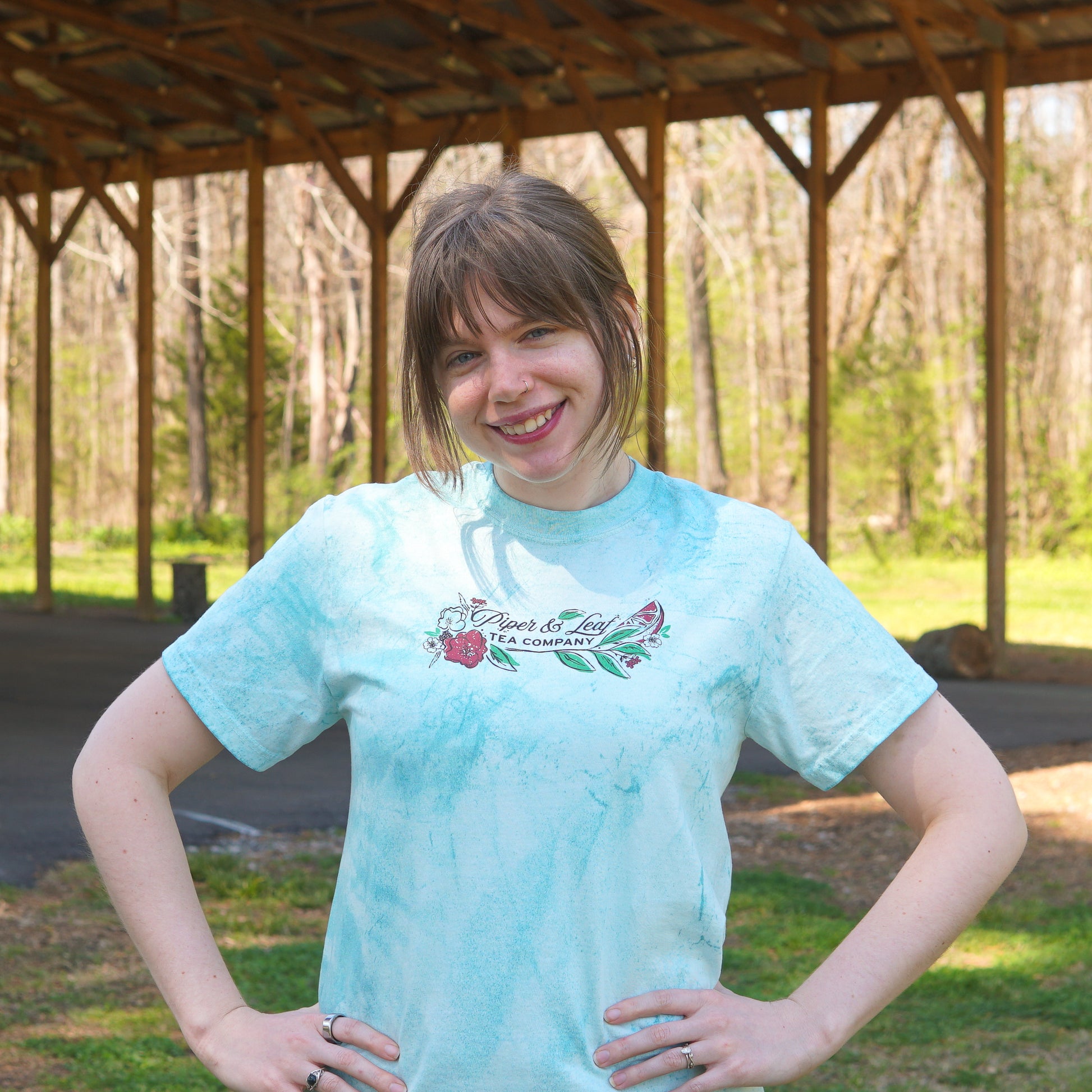 Woman with light brown hair and bangs stands smiling outdoors under a wooden shelter, wearing a light blue "Berries in Bloom Short Sleeve T-shirt" by Piper and Leaf Tea Co., hands on hips.