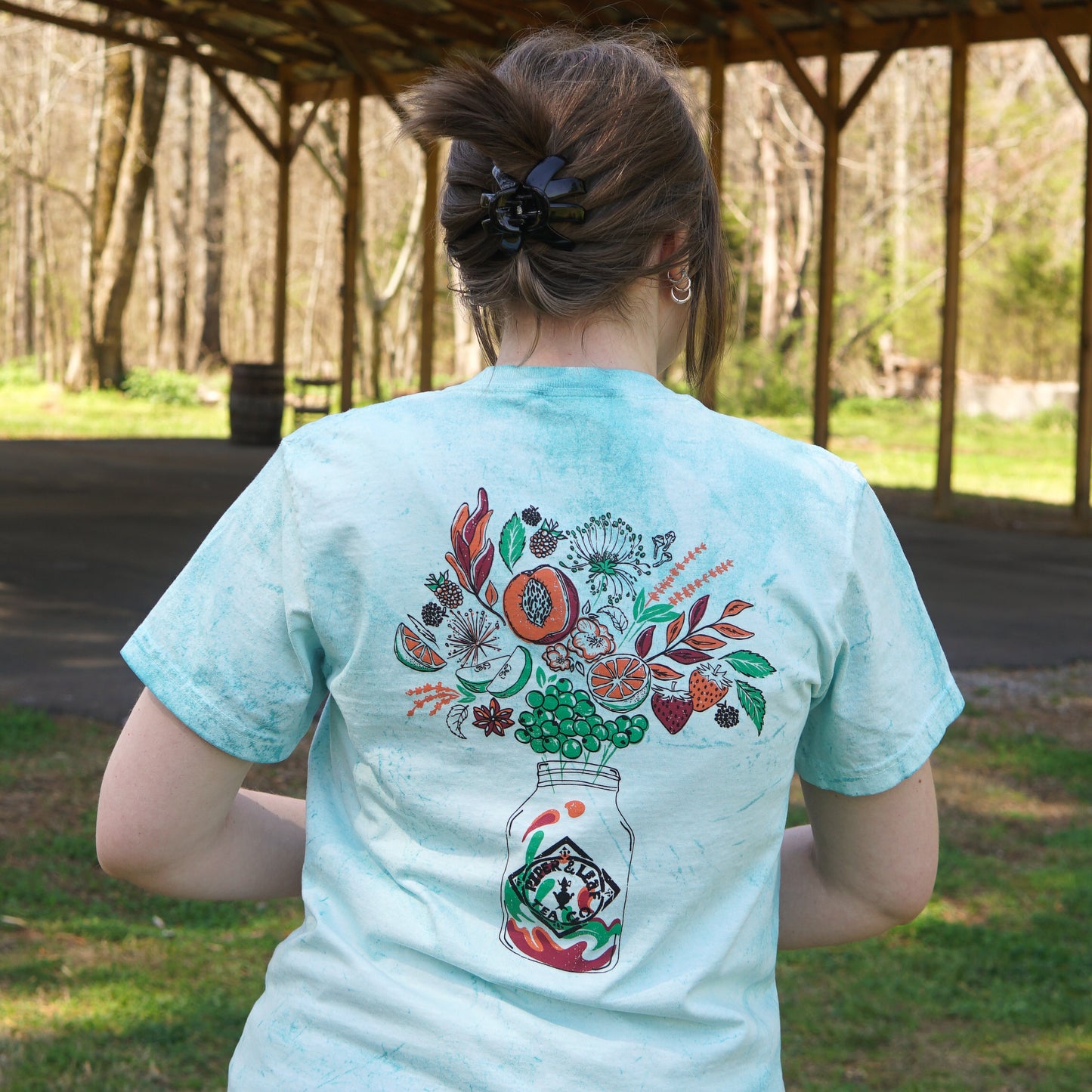 A person with light brown hair in a clip wears a Piper and Leaf Tea Co. Berries in Bloom Short Sleeve T-shirt with a floral mason jar design on the back, standing outside under a wooden shelter.