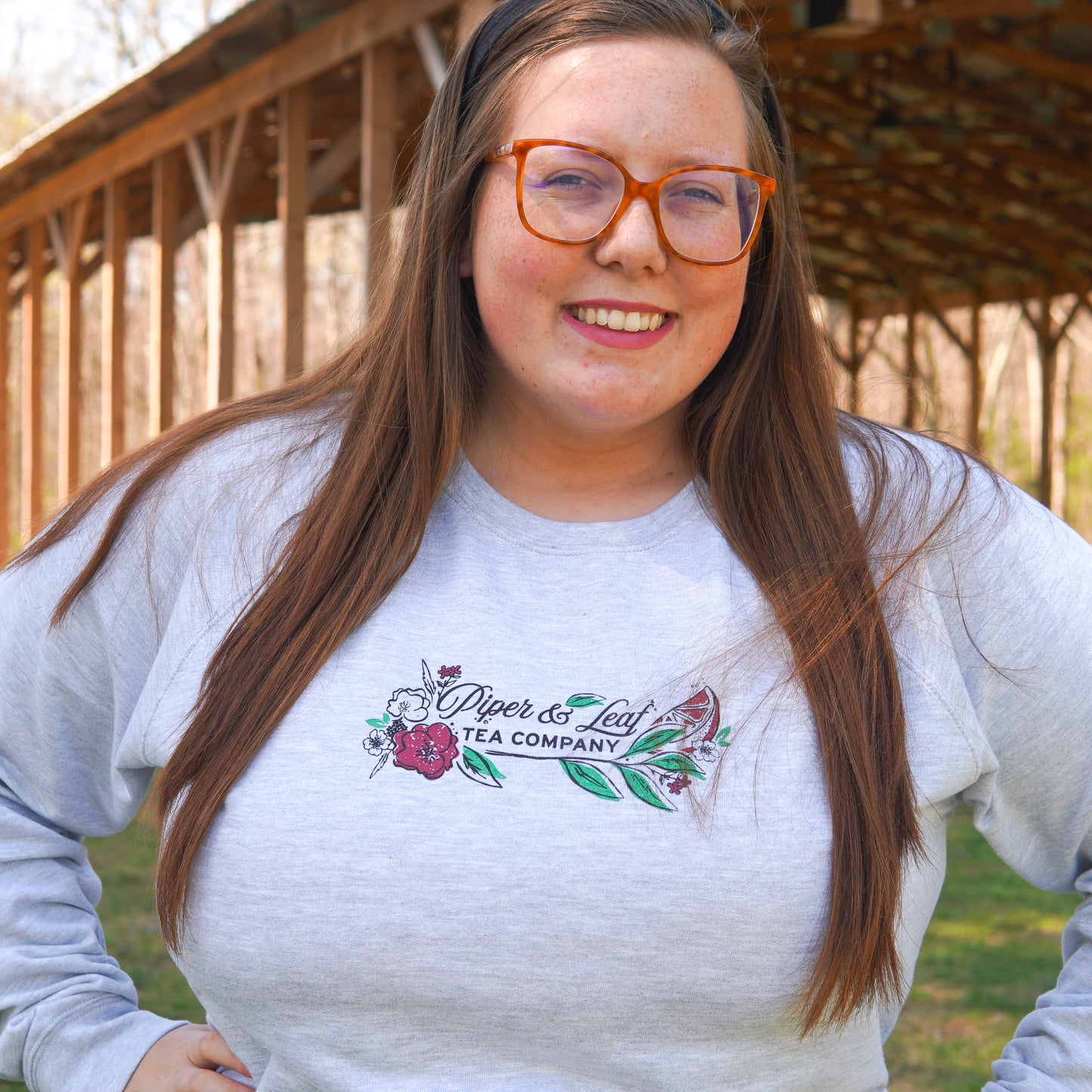 A woman with long brown hair and glasses wears a light gray Berries in Bloom Sweatshirt by Piper and Leaf Tea Co. made from Grey French Terry, standing outdoors near a wooden structure.