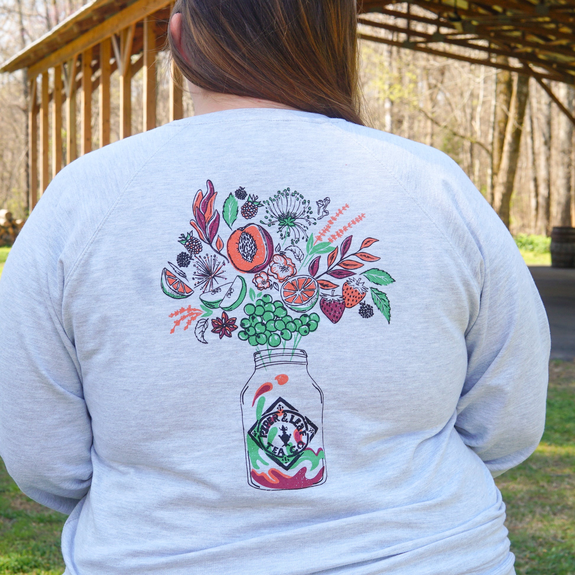A person wears the Piper and Leaf Tea Co. Berries in Bloom Sweatshirt, featuring a vibrant mason jar graphic with fruits, herbs, and flowers on the back, while standing outdoors near a wooden structure.