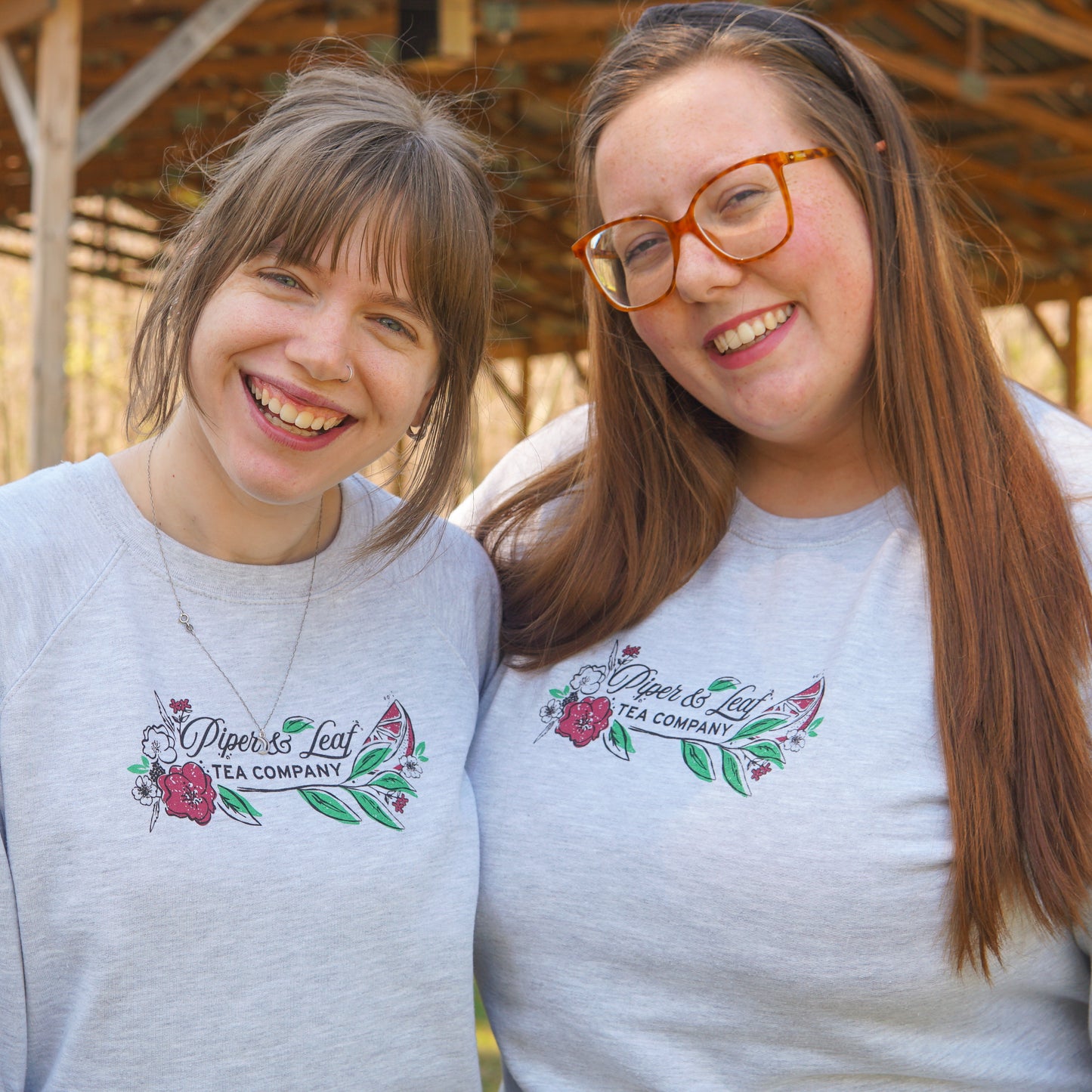 Two people stand side by side, smiling at the camera in matching Piper and Leaf Tea Co. "Berries in Bloom" sweatshirts featuring a floral logo. A wooden structure can be seen in the background.
