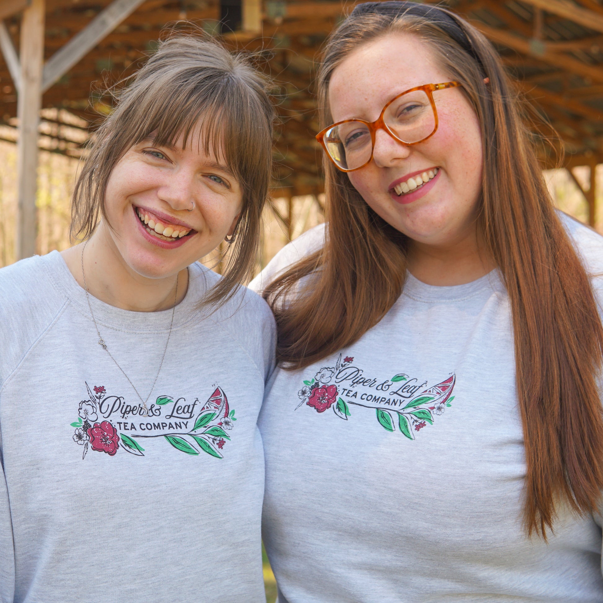 Two people stand side by side, smiling at the camera in matching Piper and Leaf Tea Co. "Berries in Bloom" sweatshirts featuring a floral logo. A wooden structure can be seen in the background.