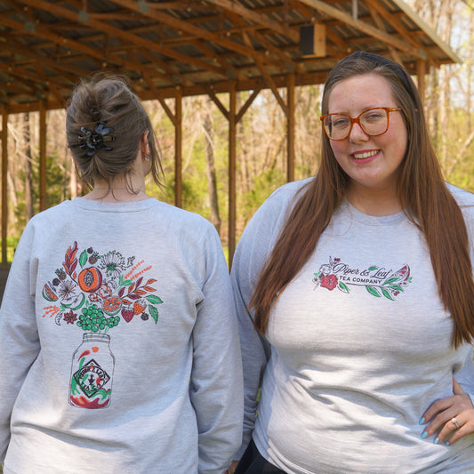 Two women wear matching light gray "Berries in Bloom Sweatshirt" by Piper and Leaf Tea Co.; one faces forward, the other shows a floral mason jar on the back. Both stand outdoors beneath a wooden structure.