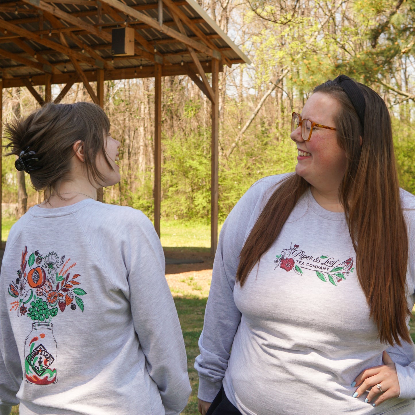 Two women wear matching Piper and Leaf Tea Co. Berries in Bloom Sweatshirts, featuring floral designs and text on light gray lightweight fabric, as they smile at each other outdoors near a wooden shelter.