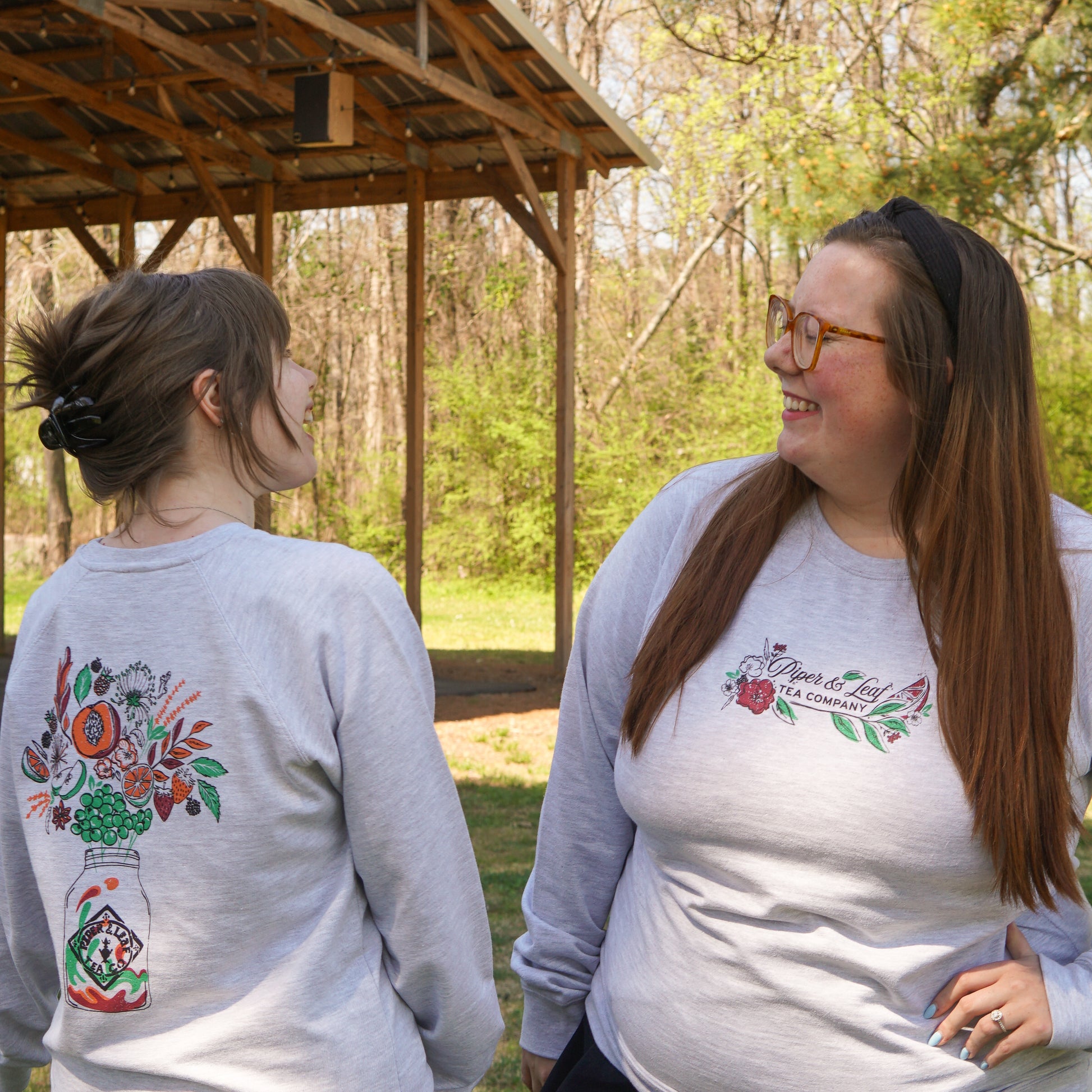 Two women wear matching Piper and Leaf Tea Co. Berries in Bloom Sweatshirts, featuring floral designs and text on light gray lightweight fabric, as they smile at each other outdoors near a wooden shelter.