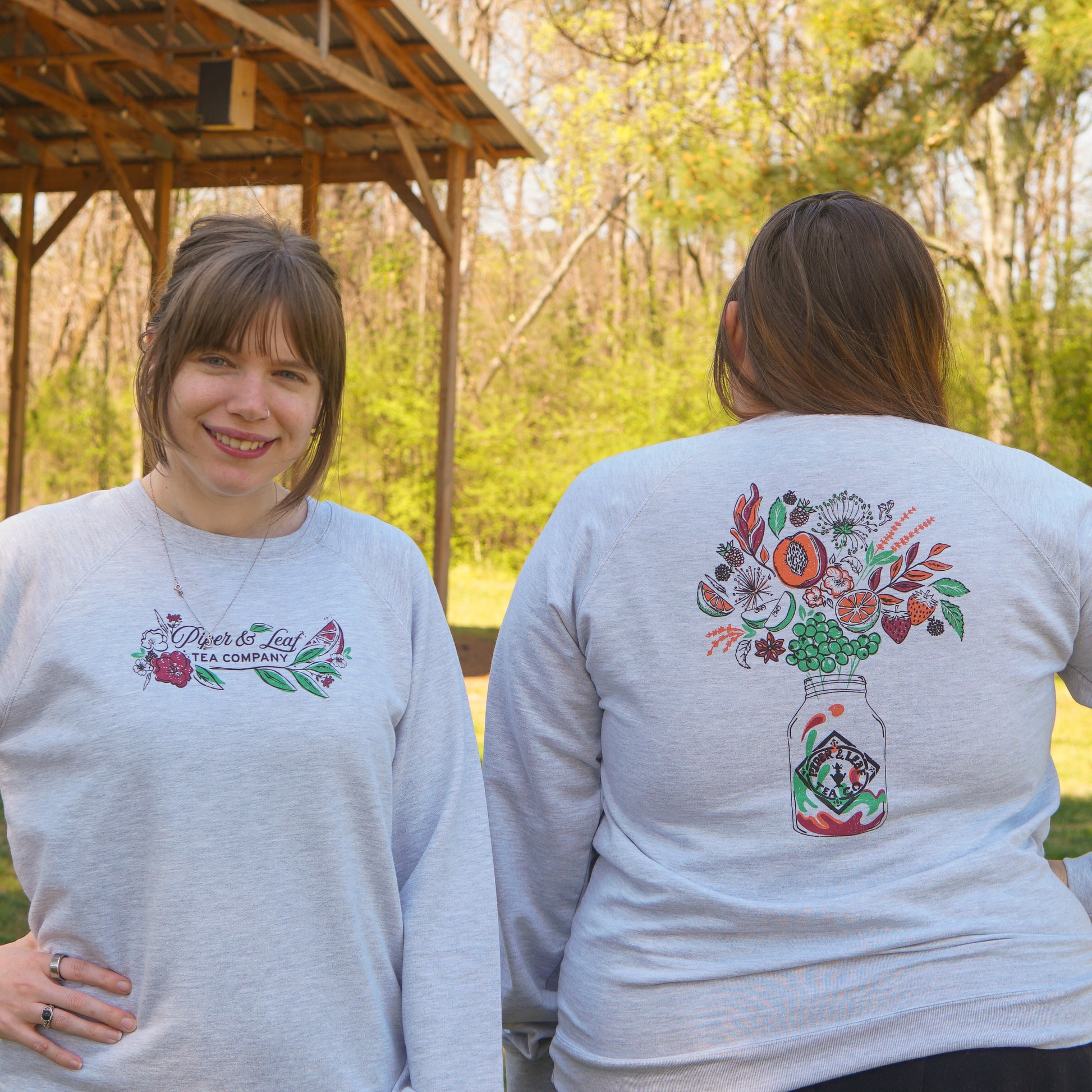 Two women stand outdoors in Piper and Leaf Tea Co.'s Berries in Bloom Sweatshirts; one faces forward, while the other reveals the back mason jar and floral design.