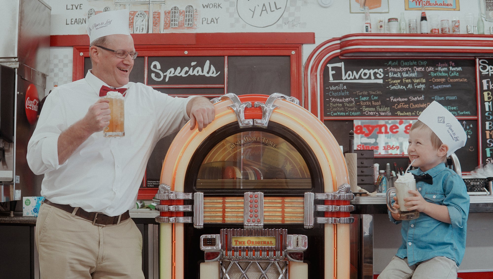 A man and a boy smile at each other, holding large mugs of Piper and Leaf Tea Co.'s Root Brew beside a vintage jukebox in a retro diner.